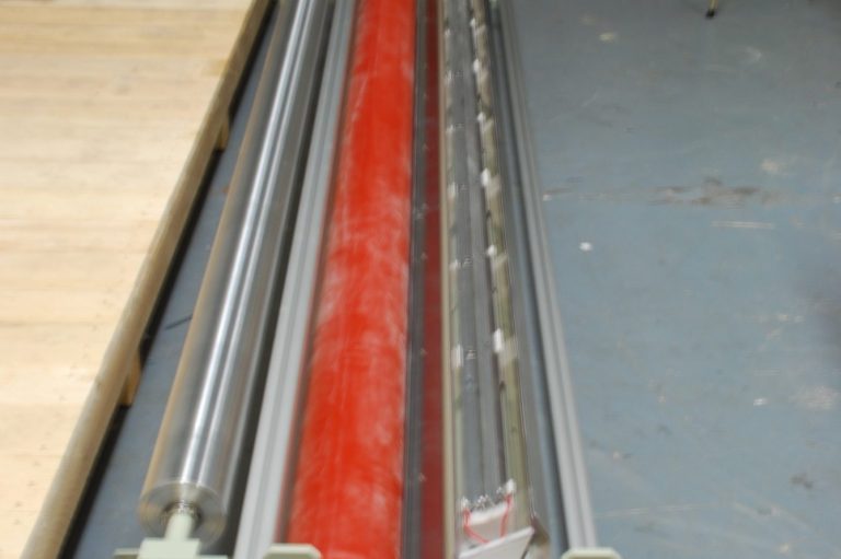 A covered roll corona treater with a red silicon sleeve coated roller and metal electrodes.